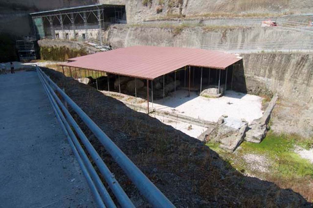 Villa dei Papiri, Herculaneum. July 2010.
Looking north-west towards area of collapsed “large or monumental structure”, possibly coinciding with the western entrance of the villa overlooking the sea. Photo courtesy of Michael Binns.
At the front of the structure is a rectangular pool parallel to the ancient coastline (the white shadowed area at the front centre in this photo).
A room of uncertain function juts out along the side of the pool. To the side of this room is a small stairway and ramp leading to the ancient beach.
See Guidobaldi, M. P. The Ivory Tripods. (p.114-5 of Buried by Vesuvius, the Villa dei Papiri at Herculaneum, edited by Kenneth Lapatin).
According to Camardo –
“The most important achievement of the open-air excavation (of 1996) was the discovery of the monumental structure with the same orientation as the Villa but situated on a large terrace some ten metres below its piano nobile, and jutting out noticeably farther towards the sea.
This was only partially unearthed because it had completely collapsed under the weight of the pyroclastic debris that had buried it.
The collapsed part was recovered whole, since the structure had not been cut into by the Bourbon-era tunnels.
A monumental hall (see fig. d in plan (14.1) on p.114) is fairly well preserved: a series of brick pilasters covered in stucco supported a set of large wooden beams with a flat ceiling in opus signinum (tiny broken tiles mixed with mortar).
A white marble threshold graced the broad entrance of the seaward side of the building.”
See Camardo, D. Recent excavations in the Villa dei Papiri 1990’s – 2008. (p.105-113 of Buried by Vesuvius, the Villa dei Papiri at Herculaneum, edited by Kenneth Lapatin).